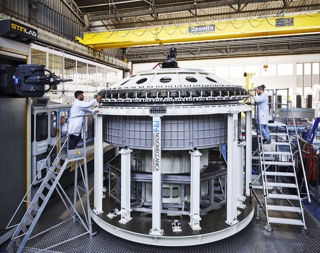 Two people wearing lab coats with the Thales Alenia Space logo guide stand on moveable staircases while working on assembling components of Axiom Station's Hab One (Ax-H1).