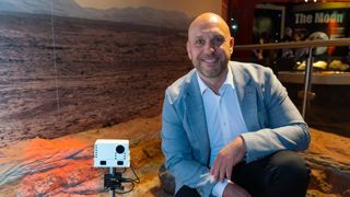 A man kneels to the right of a space rover on a replica of Mars, The man is smiling and wearing a grey jacket and white shirt.