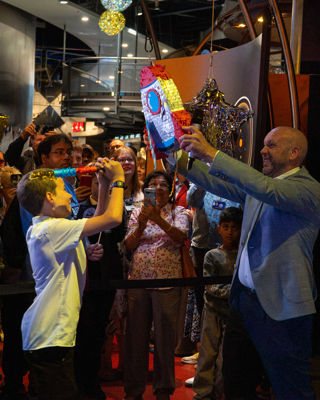 A school child attempts to hit a rocket-shaped pinata that is being held by a man. People are watching in the background.
