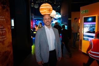 A man stands in a gallery smiling. He is wearing a grey jacket and white shirt. Behind him are screens and people. Above him is a replica planet. 